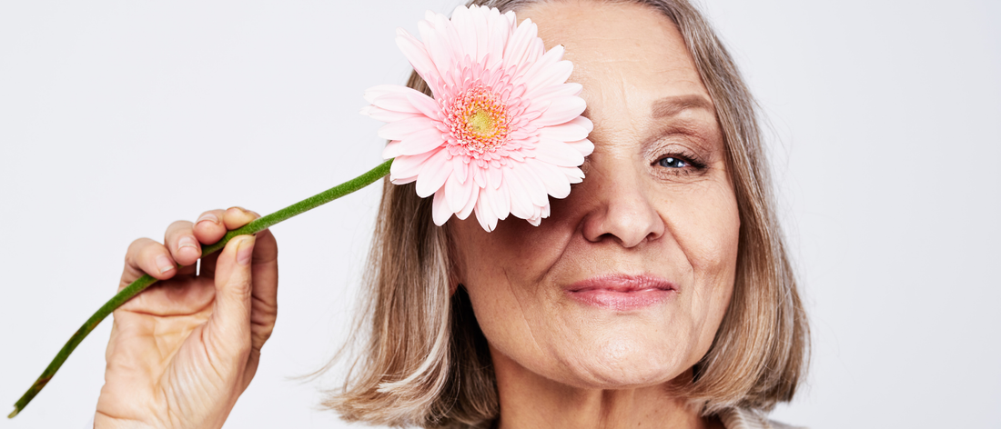 Mujer sonriente en la menopausia sosteniendo una flor rosa sobre un ojo, mostrando su piel radiante y su bonito cabello tras tomar colágeno bebible