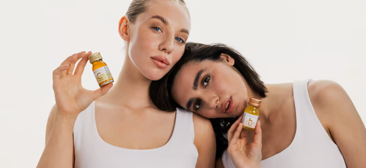 La morena y la rubia se apoyan una en otra sosteniendo en sus manos colágeno Primabiotic y ácido hialurónico en frascos.
