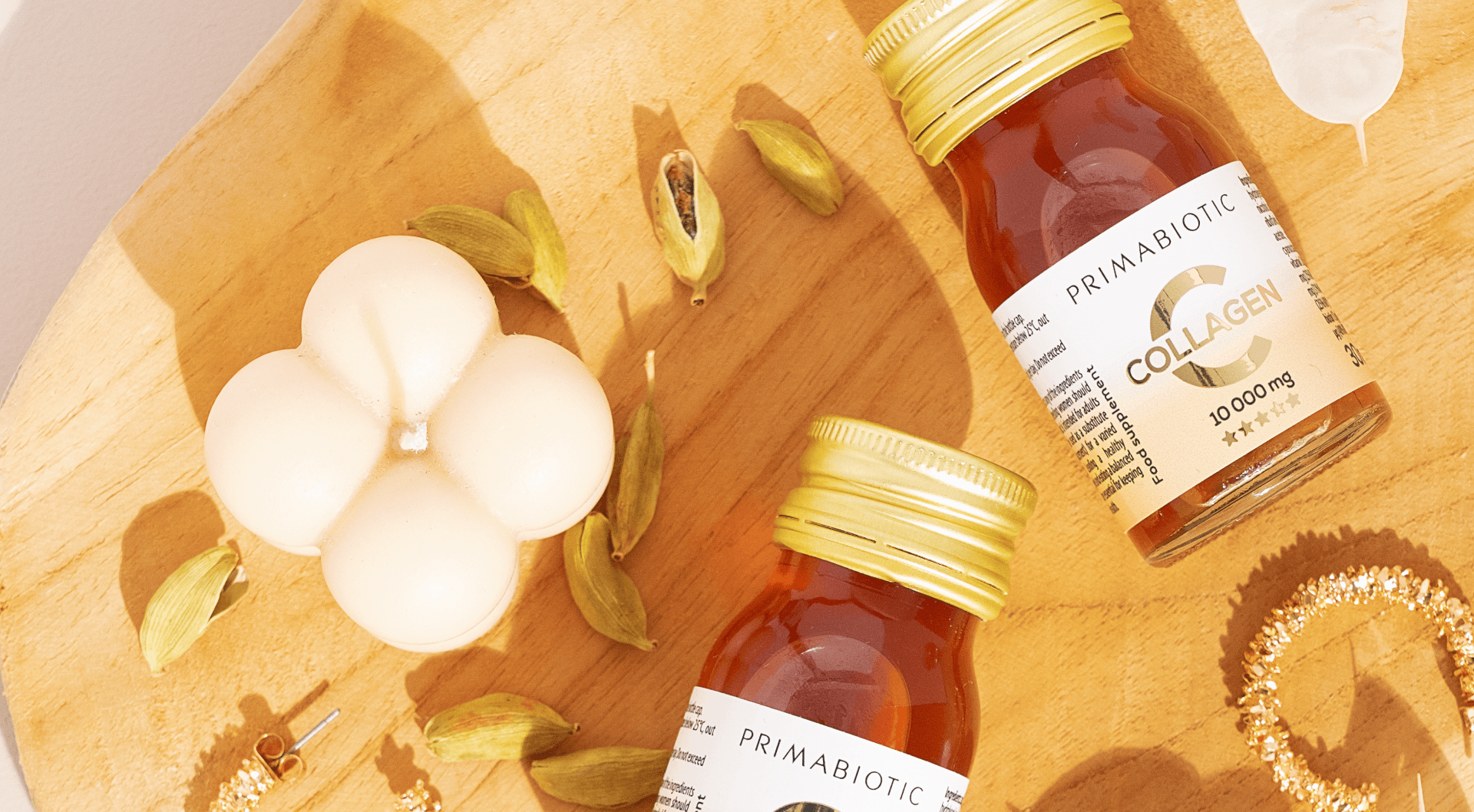 Dos botellitas de suplemento Primabiotic Collagen colocadas sobre una bandeja de madera junto a una vela decorativa, especias y accesorios dorados
