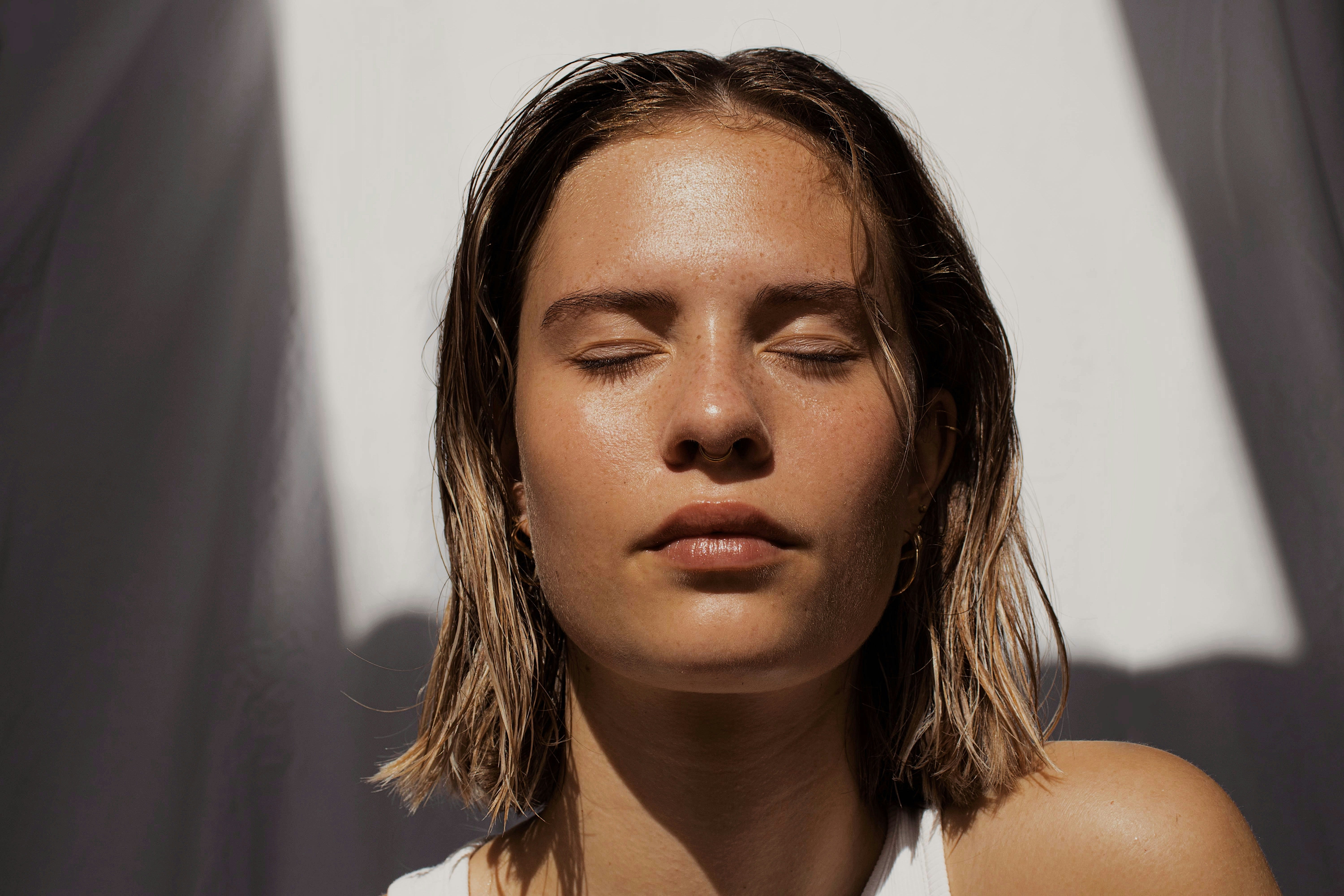 Retrato de mujer joven con cabello rubio corto y ojos cerrados, disfrutando de la luz solar que resalta su piel saludable y luminosa, imagen beauty
