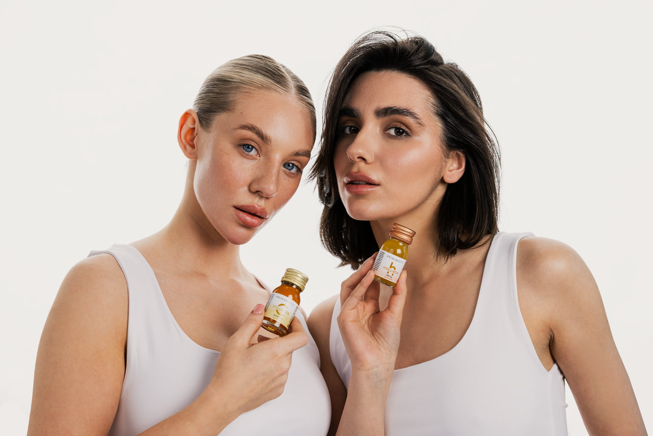 Dos mujeres jóvenes posan con suplementos Primabiotic: una sostiene botellita de colágeno y la otra de ácido hialurónico