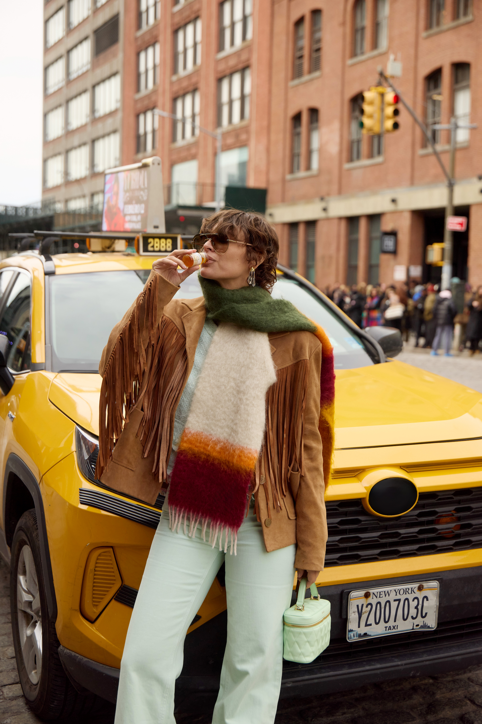 Mujer con bufanda de colores bebe botellita de Primabiotic junto a taxi amarillo en la ciudad